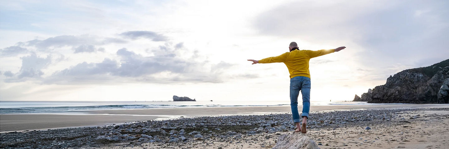 A man in a yellow sweater and jeans with arms outstretched balancing on a rock on the beach