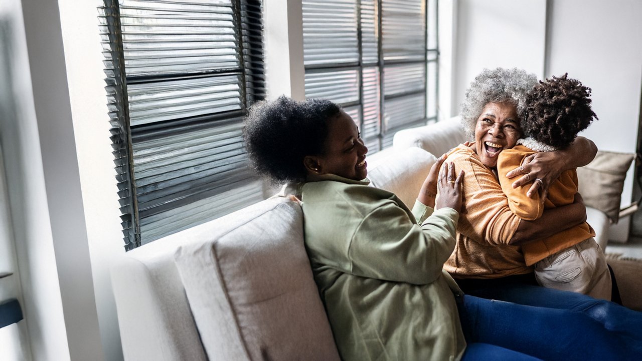 Smmiling grandparent sitting beside her child on the couch being hugged by grandchild