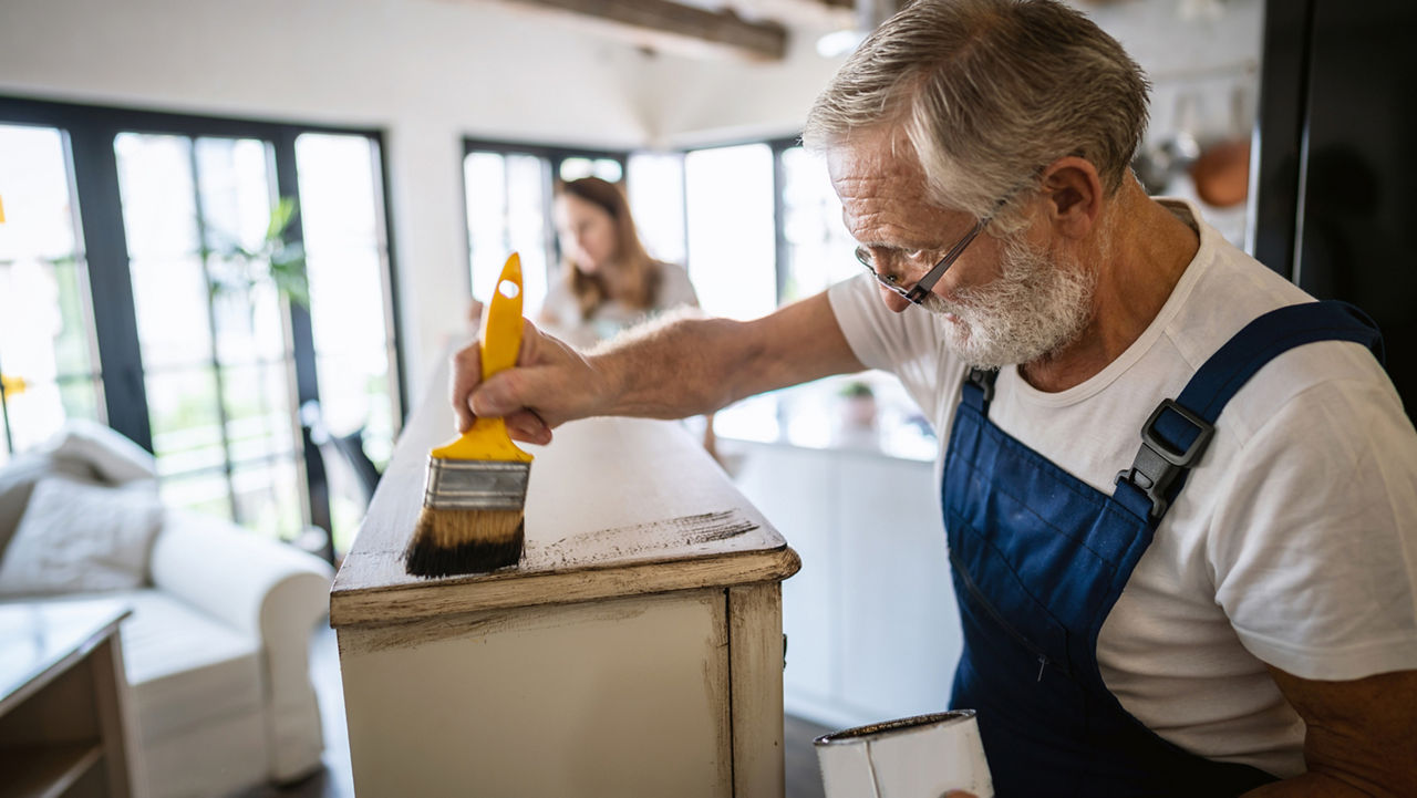 An older man wearing dungarees, wearing glasses and painting a wooden cabinet