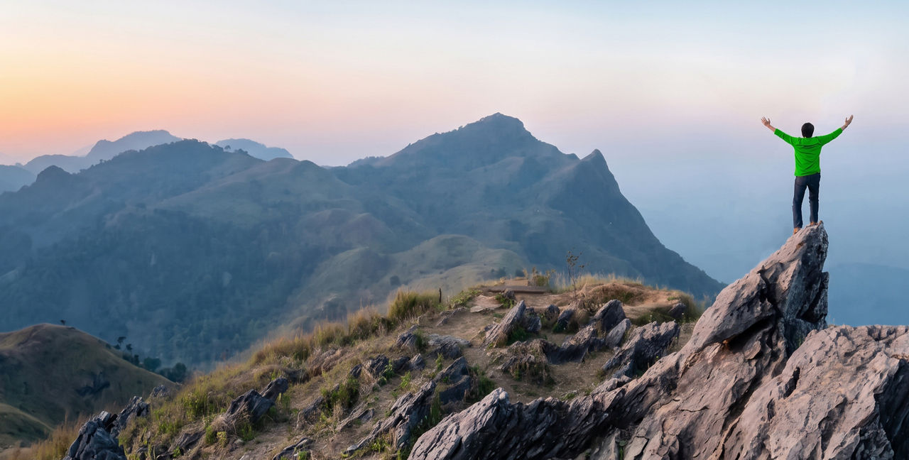 Person standing on a rocky mountain outcrop with arms raised, overlooking a mountain landscape.